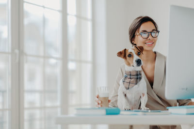 Smiling young woman with dog using computer on desk in office
