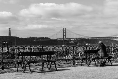 Rear view full length of man sitting on bench against april 25th bridge