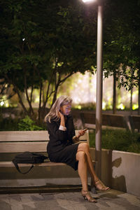 Full length of businesswoman using phone while sitting on bench at park