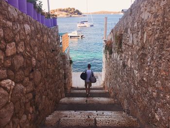 Rear view of person carrying luggage on staircase amidst stone wall against sea