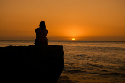 Rear view of woman sitting on rock by sea against sky during sunset