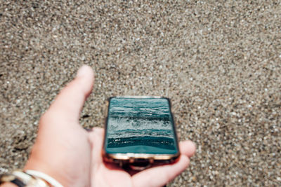 Close-up of hand holding smart phone reflected sea against beach