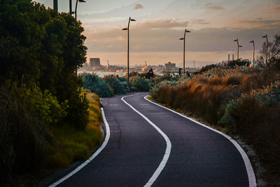 Empty road by trees against sky during sunset