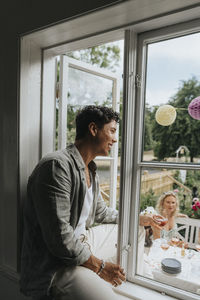 Smiling young man sitting on window sill while looking at friends celebrating party at cafe