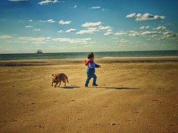 Full length of boy playing on beach with dog against sky