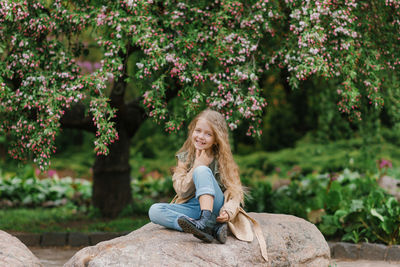 Caucasian cute baby girl with long wavy blonde hair in a trench coat sits on a stone in spring 