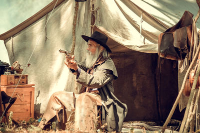 Bearded man aiming gun while sitting against tent