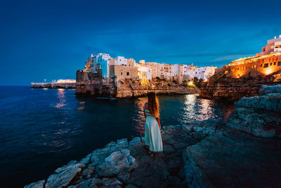 Illuminated buildings by sea against blue sky