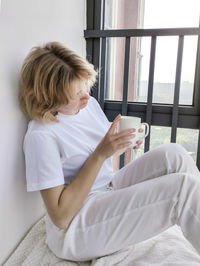 Attractive young woman sitting on the windowsill and holding a cup