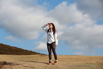 Full length of woman shielding eyes while standing on field against sky