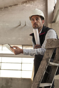 Male engineer with digital tablet and blueprint contemplating while standing in building at site