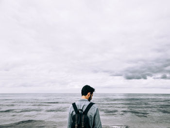 Rear view of teenage boy with backpack standing by sea against cloudy sky