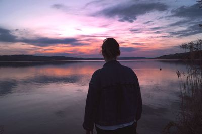 Rear view of woman standing by lake against sky during sunset