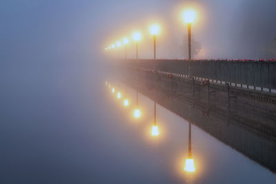 Illuminated street light by river against sky at night