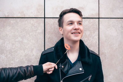 Cropped image of hand holding kiss prop by thoughtful smiling young man against wall
