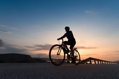 Man riding bicycle on street against sky during sunset