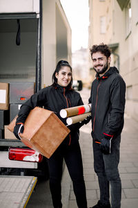 Portrait of confident young delivery man and woman with packages standing against truck in city