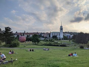 People relaxing on field by buildings against sky