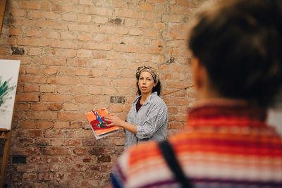 Instructor showing painting to artist while standing against brick wall at studio