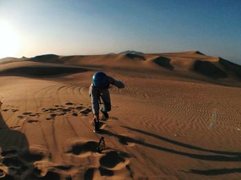 Boy reaching for eyewear on sand dune in desert against sky