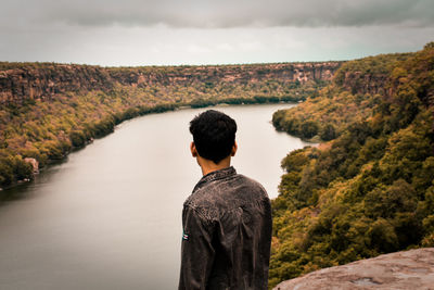 Side view of young man looking at view while standing on mountain over river against cloudy sky