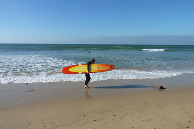 Side view of man holding surfboard while walking on sea shore against sky