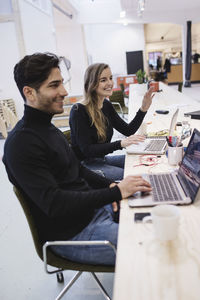 Cheerful young woman and man looking away while sitting at desk in office