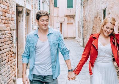 Smiling young couple holding hands while walking on footpath against buildings in city