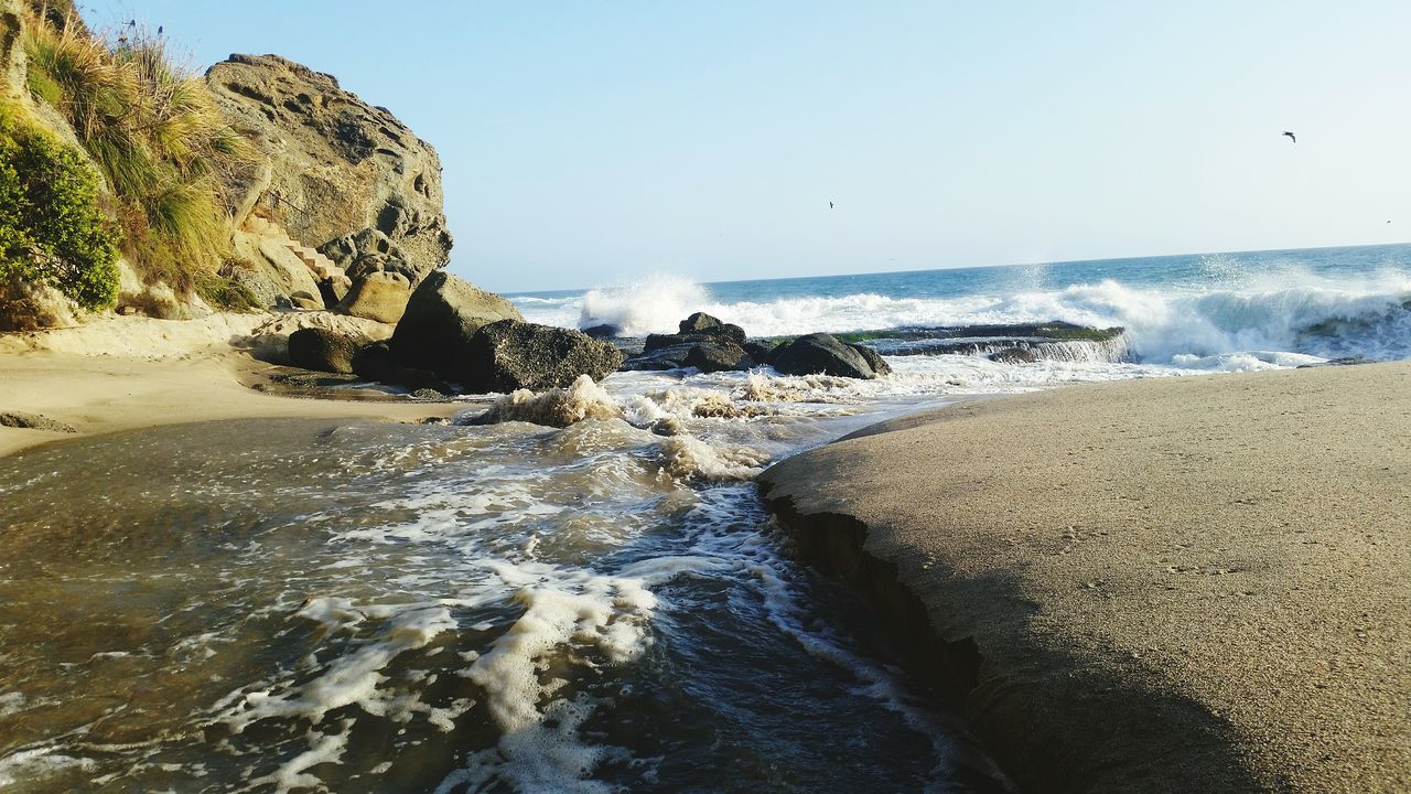 sea, water, beach, clear sky, wave, surf, scenics, beauty in nature, horizon over water, shore, rock - object, nature, motion, tranquil scene, rock formation, tranquility, splashing, idyllic, sand, copy space