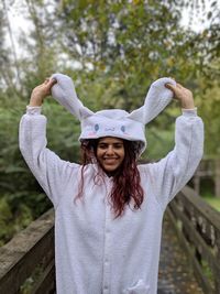 Portrait of young woman wearing white warm clothing while standing on footbridge against trees