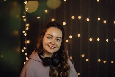 Young smiling woman with headphones, sitting near glowing christmas tree with bokeh, evening