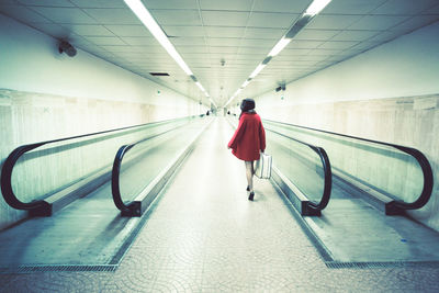 Rear view of woman with luggage walking on pathway amidst moving walkway in illuminated subway