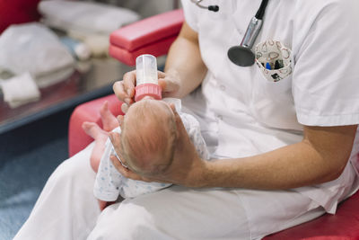 Doctors sitting on armchairs and feeding newborns from bottles holding heads in room with equipment in hospital