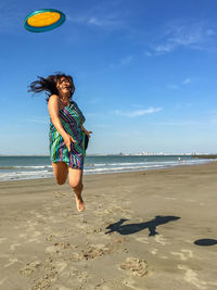 Full length of woman jumping while throwing plastic disc at beach against sky during summer