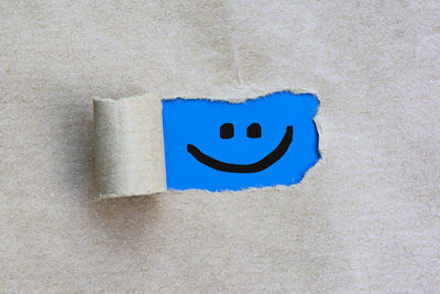Happy smiley face on torn hole in the sheet of brown paper on blue background.