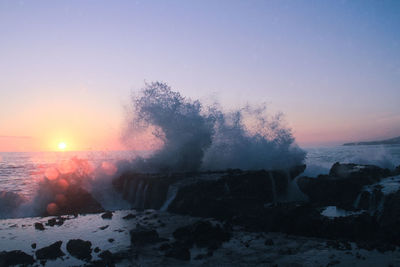 Sea waves splashing on rocks against sky during sunset