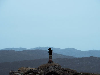 Man standing on cliff in front of mountains against clear sky