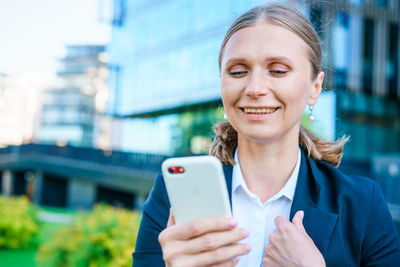 Successful female banker using smart phone outdoors while standing near office