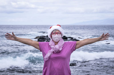 Portrait of senior woman wearing mask with arms outstretched standing against sea