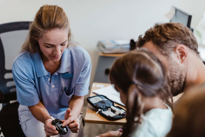 Female pediatrician showing glaucometer to girl sitting with father in hospital