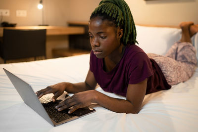 Full body of young ethnic female lying on bed and browsing netbook while working remotely in modern bedroom at home