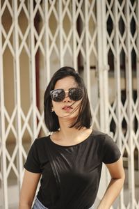 Portrait of young woman wearing sunglasses standing against fence