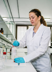 Focused female scientist in white uniform and latex gloves mixing chemical liquids in flask while working in modern light lab