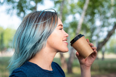 Blue haired woman drinks take away coffee outdoors on nature background.