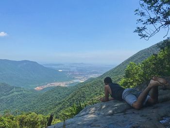 Man sitting on retaining wall by mountains against clear sky