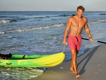 Young fit millennial man pulling kayak out of ocean.