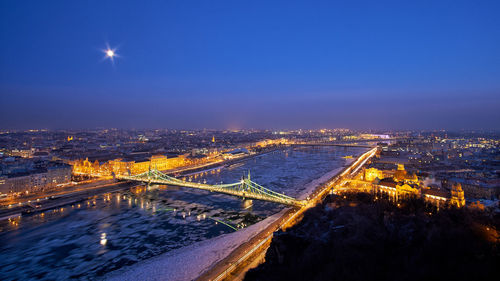 Bridge over river in illuminated city against sky