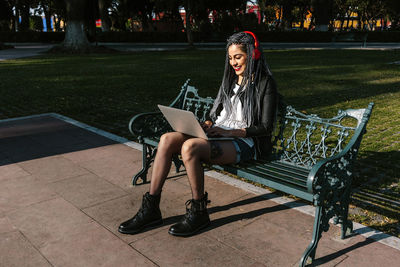 Content latin american female in trendy apparel and headphones sitting on bench with netbook during video chat in sunny park