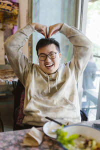 Happy young man making heart shape with hands while sitting at restaurant