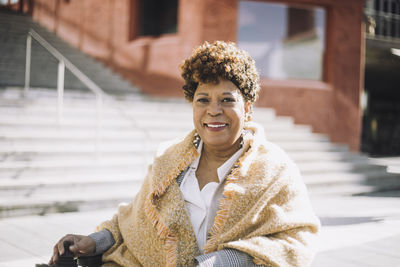 Portrait of smiling senior woman with curly hair sitting on steps during sunny day
